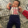 baby, infant, clothing, blanket, pattern, lying_down, flexing, cute, blue_pants, blue_slippers, red_shirt, text_on_clothing, indoor, bed, fabric, person, child, face, small_hands, cute_expression