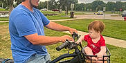 Zayden is registered to the contest to win money with this photo: man, baby, electric_bike, blue_shirt, red_clothing, basket, grass, park, trees, side_view, outdoor, daytime, cap, jeans, curious, handlebars, wheel, sky, cloudy, people