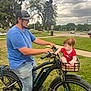baby, basket, blue_shirt, cap, cloudy, curious, daytime, electric_bike, grass, handlebars, jeans, man, outdoor, park, people, red_clothing, side_view, sky, trees, wheel