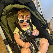 Zayden is registered to the contest to win money with this photo: baby, child, sunglasses, stroller, infant, plaid, romper, clothing, seatbelt, indoor, floor, wood_floor, person, cute, hand, legs, white_socks, hair, face, comfort