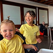 Ishaq participe au concours pour gagner de l'argent avec cette photo : child, toddler, yellow_shirt, smile, laughing, indoor, high_chair, hair_tie, wooden_beams, table, person, happy, casual_clothing, playful, sitting, family, background, blurred, friendship, joy