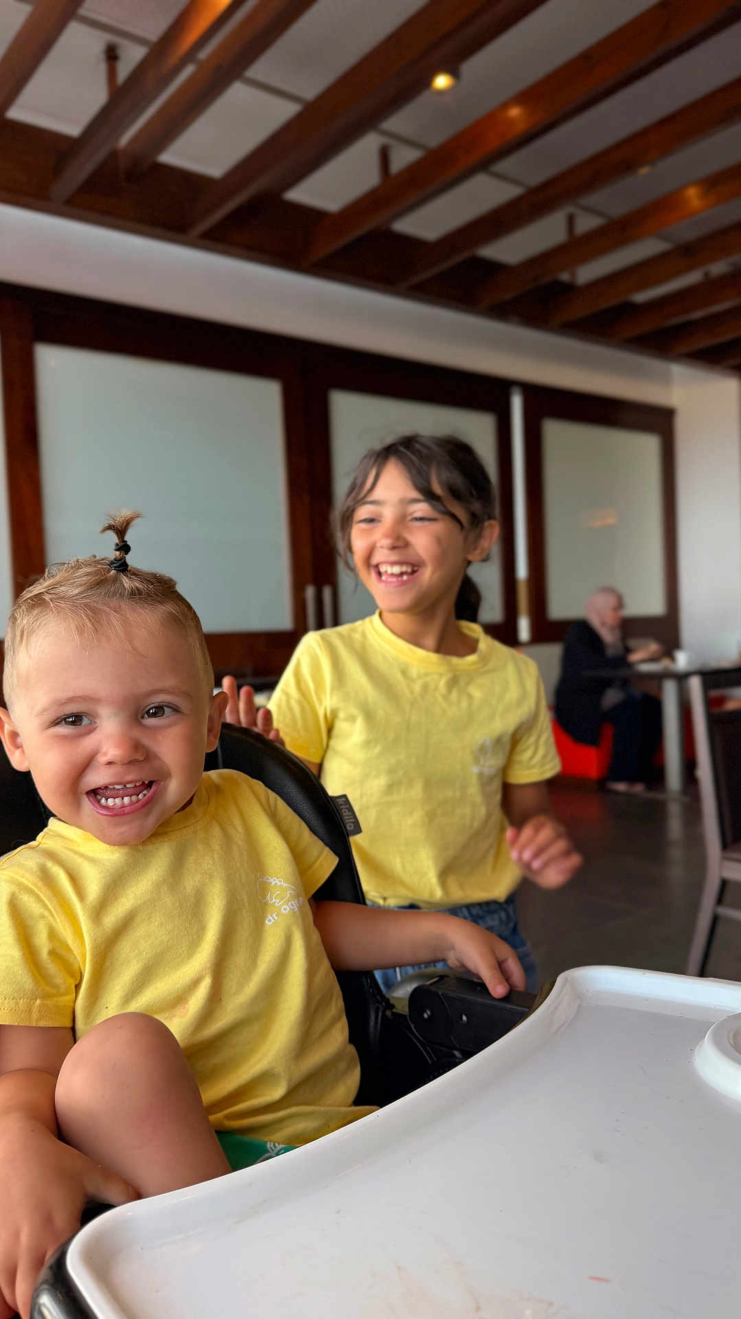 Ishaq participe au concours pour gagner de l'argent avec cette photo : child, toddler, yellow_shirt, smile, laughing, indoor, high_chair, hair_tie, wooden_beams, table, person, happy, casual_clothing, playful, sitting, family, background, blurred, friendship, joy