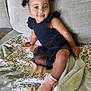 toddler, child, smiling, indoor, couch, blanket, floral_pattern, black_dress, pink_shoes, curly_hair, pigtails, happy, person, sitting, cute, portrait, cozy, home, soft_lighting, fashion