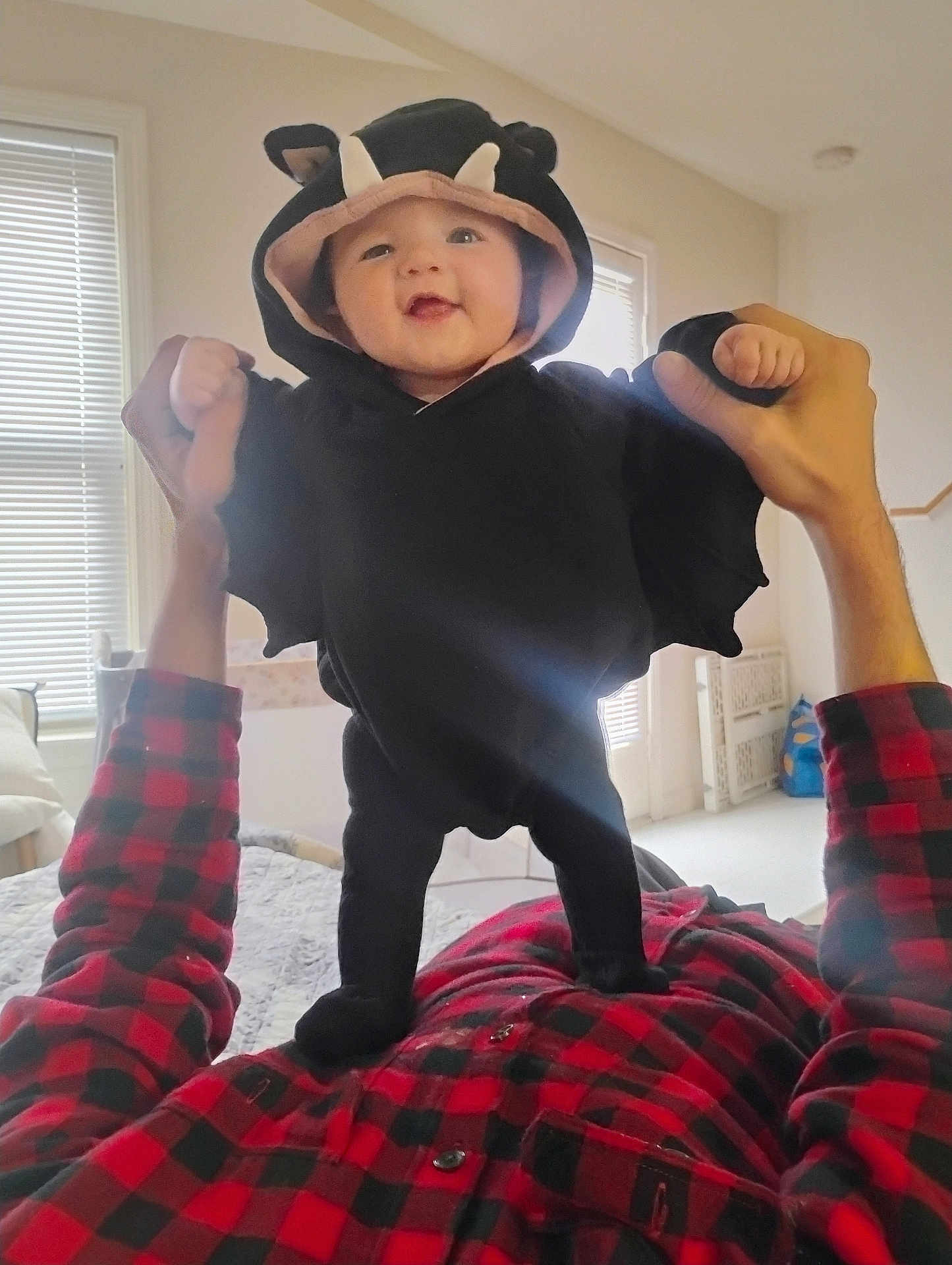 Raelynn joined the competition — help win amazing prizes! baby, child, costume, bat, black_clothing, hood, smile, hands, adult, plaid_shirt, indoor, window_blinds, bed, blanket, light, person, cute, fun, playful, family