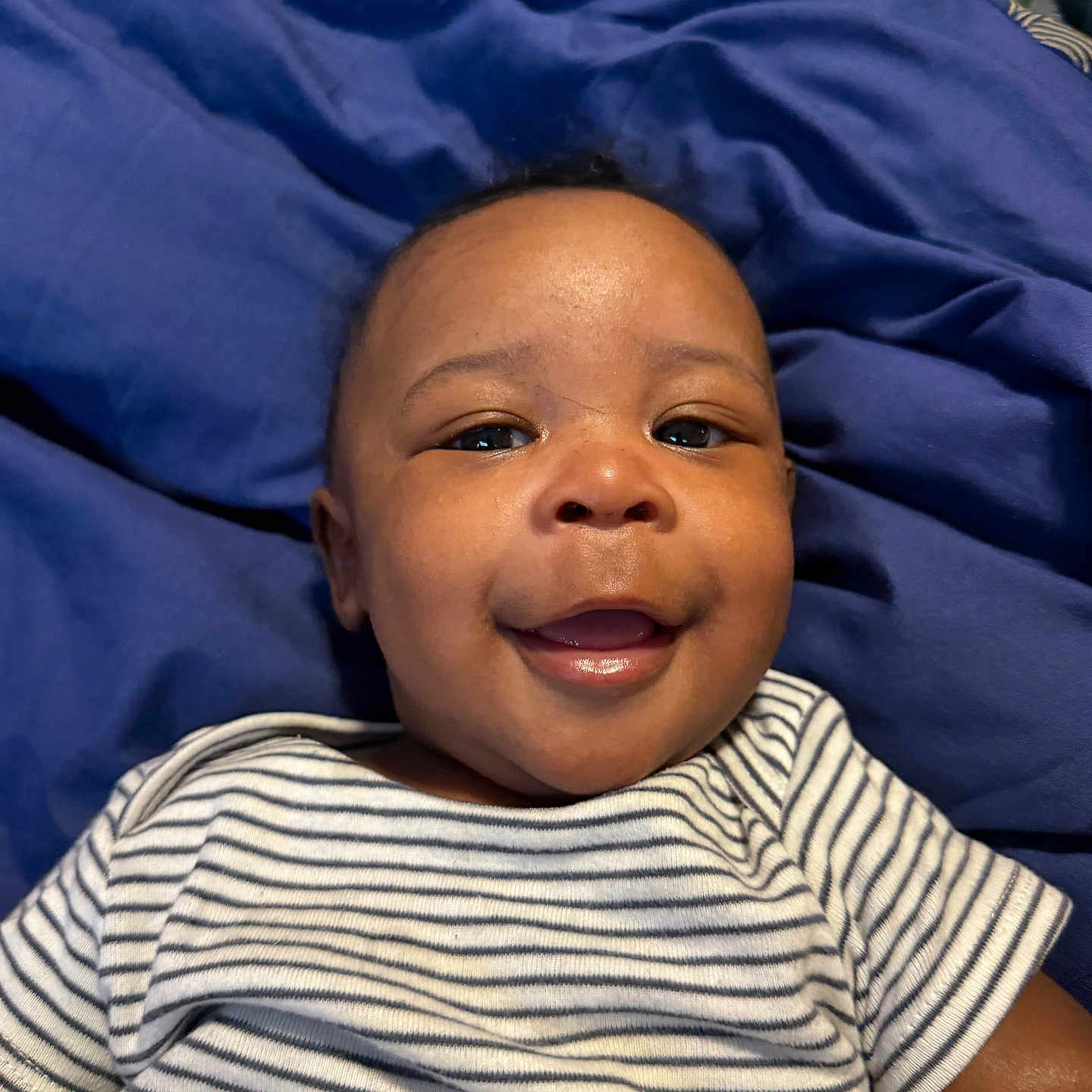 Auren is registered to the contest to win money with this photo: baby, child, smiling, face, portrait, striped_shirt, clothing, lying_down, blue_blanket, happy, infant, cute, person, young, skin, head, eyes, mouth, indoors, baby_boy
