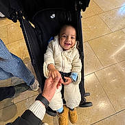 Emaan joined the competition — help win amazing prizes! toddler, child, stroller, hand_holding, smile, white_snowsuit, shoes, floor_tiles, indoor, adult_hand, casual_clothing, joyful, baby, person, footwear, black_stroller, happy, candid, human_interaction, winter_clothing