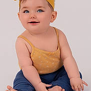Elsie is registered to the contest to win money with this photo: baby, child, smiling, headband, yellow, blue_pants, sitting, happy, cute, portrait, indoors, infant, skin, face, hands, feet, clothing, studio, young, person
