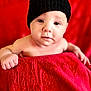 baby, infant, face, hat, black_hat, red_blanket, red_background, skin, eyes, hand, cute, portrait, child, young, looking, expression, indoors, soft_texture, warm_colors, cozy