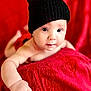 baby, child, infant, black_hat, red_blanket, soft_texture, close_up, cute, portrait, skin, hand, face, eyes, lying_down, warm_colors, indoor, cozy, knitwear, expression, adorable