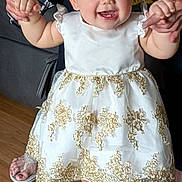 Nevaeh is registered to the contest to win money with this photo: baby, toddler, child, smiling, dress, white_dress, gold_embroidery, holding_hands, adult_hands, person, face, teeth, shoes, socks, hardwood_floor, living_room, sofa, portrait, cute, celebration