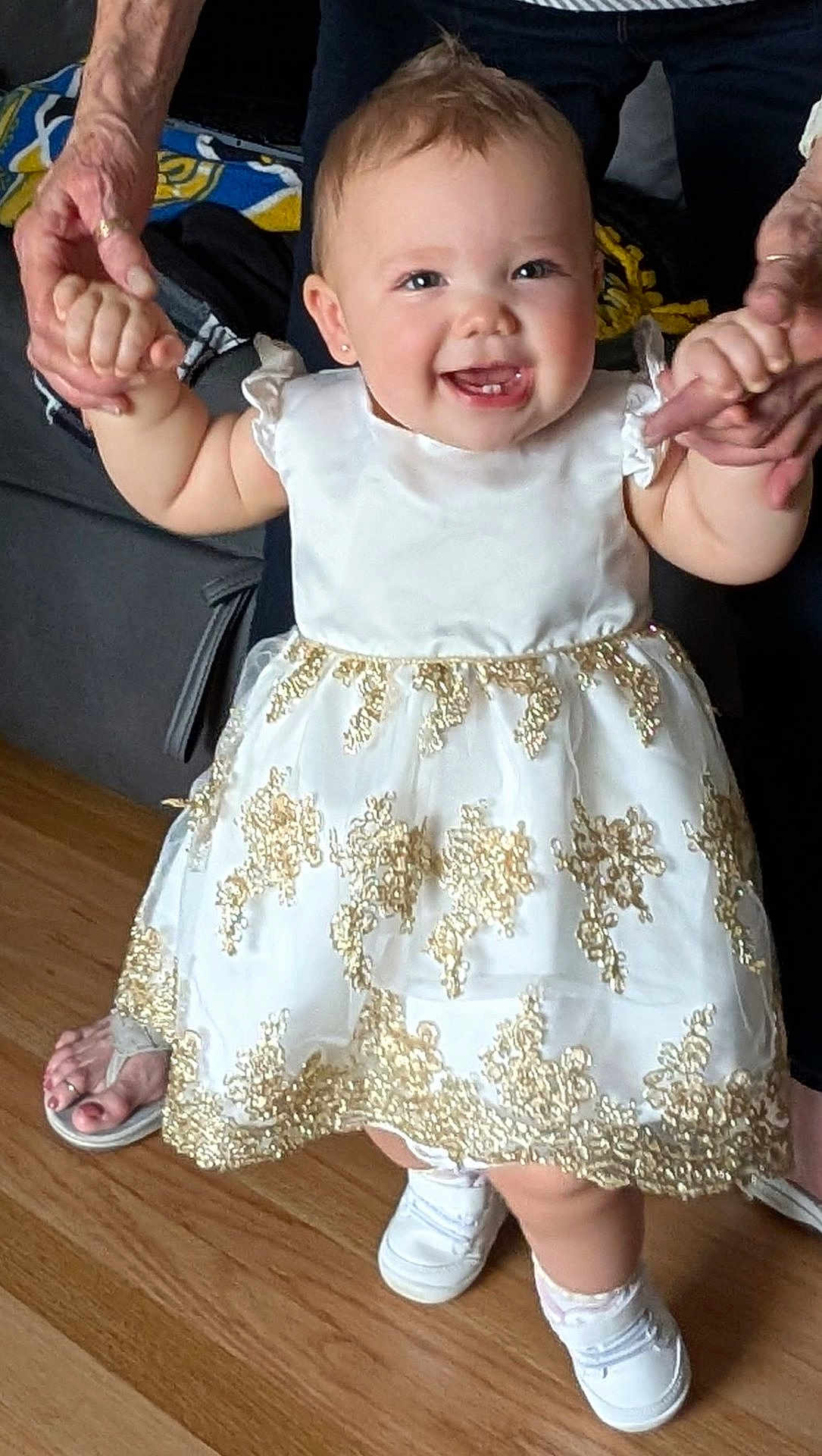Nevaeh is registered to the contest to win money with this photo: baby, toddler, child, smiling, dress, white_dress, gold_embroidery, holding_hands, adult_hands, person, face, teeth, shoes, socks, hardwood_floor, living_room, sofa, portrait, cute, celebration