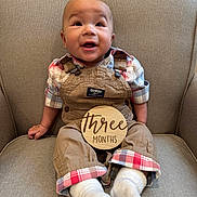 Gracen joined the competition — help win amazing prizes! baby, infant, child, plaid_shirt, overalls, socks, wooden_sign, armchair, smiling, happy, seated, indoors, cute, person, face, clothing, fabric, furniture, portrait, three_months