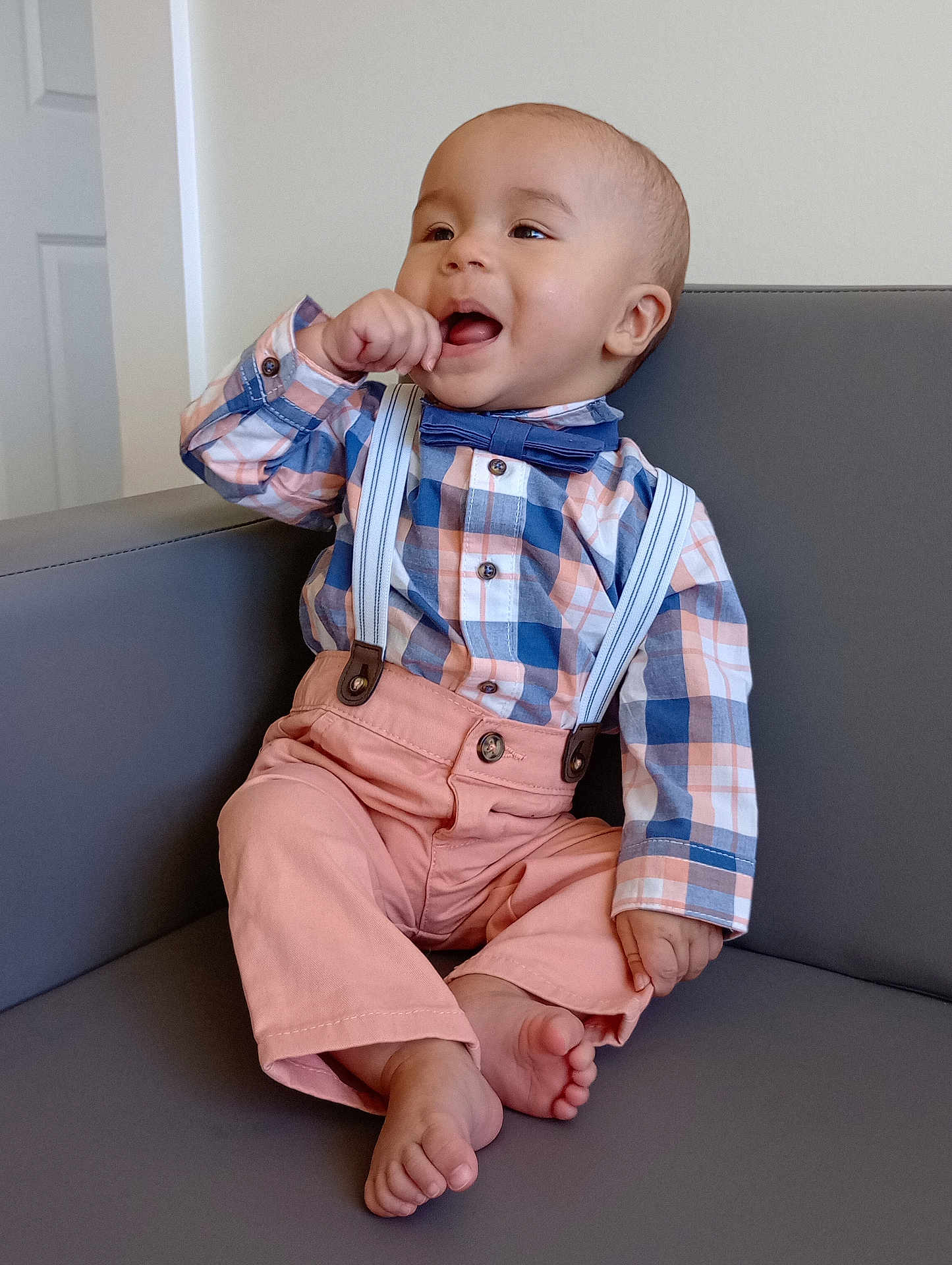 Gracen is registered to the contest to win money with this photo: baby, child, boy, smiling, plaid_shirt, bow_tie, suspenders, pink_pants, barefoot, couch, indoor, happy, cute, portrait, sitting, furniture, hand_to_mouth, casual, infant, light_background