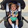 baby, child, stroller, hat, sunhat, socks, shorts, shirt, dinosaur_pattern, smiling, happy, outdoor, pavement, footwear, black_stroller, seat, sunlight, infant, person, cute