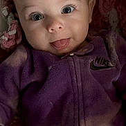 Charlotte is registered to the contest to win money with this photo: apparel, baby, blanket, child, cozy, cute, eyes, face, happy, head, indoor, infant, lying_down, nike_logo, person, portrait, purple_clothing, smiling, soft_texture, tongue_out