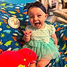 baby, child, smiling, happy, headband, dress, toy, plush, red, blue, blanket, dinosaur, pattern, sitting, indoor, cute, infant, playful, tongue_out, person