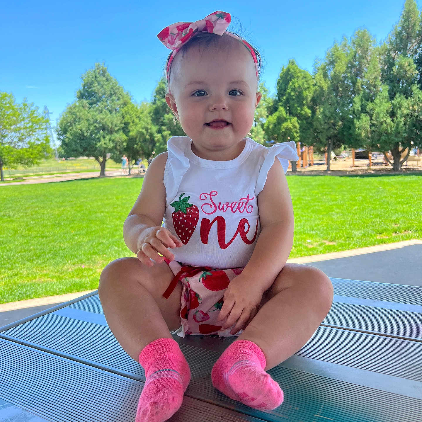 Evelynn is registered to the contest to win money with this photo: baby, child, clothing, cute, daylight, face, girl, grass, happy, headband, nature, outdoor, park, picnic_table, sitting, smiling, socks, summer, sunny, trees