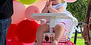 Evelynn is registered to the contest to win money with this photo: baby, child, high_chair, balloons, pink, red, green, outdoor, grass, celebration, party, smiling, clapping, headband, person, summer, daylight, decorations, toddler, feet