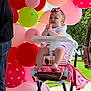 baby, child, high_chair, balloons, pink, red, green, outdoor, grass, celebration, party, smiling, clapping, headband, person, summer, daylight, decorations, toddler, feet
