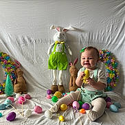 Evelynn joined the competition — help win amazing prizes! baby, child, easter_eggs, plush_bunny, wreath, colorful, smiling, sitting, decorations, toy, pastel_colors, holiday, festive, tablecloth, green_bow, white_background, cute, indoors, playful, happy