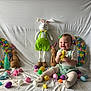 baby, child, easter_eggs, plush_bunny, wreath, colorful, smiling, sitting, decorations, toy, pastel_colors, holiday, festive, tablecloth, green_bow, white_background, cute, indoors, playful, happy