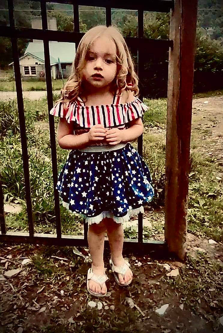 Rosalie is registered to the contest to win money with this photo: baby_toddler_clothing, blond, brown_hair, child, day_dress, face, fun, grass, happy, human_leg, knee, leisure, magenta, pattern, people_in_nature, person, plant, tints_and_shades, toddler, tree