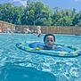 toddler, child, pool, water, inflatable_ring, swimming, summer, outdoor, sunlight, adult, fence, trees, blue_sky, recreation, leisure, vacation, fun, holiday, swimwear, relaxation