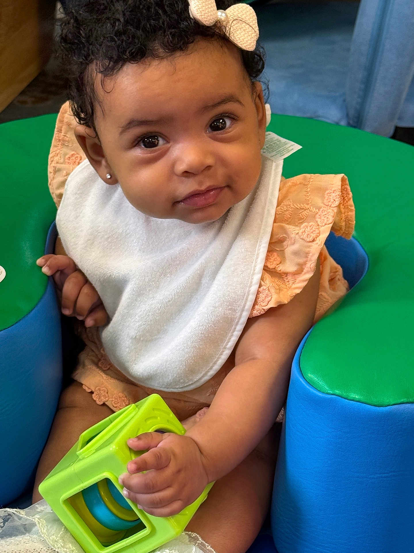 Carrera joined the competition — help win amazing prizes! baby, child, curly_hair, bow, bib, peach_dress, toy, green_toy, seat, padded_seat, hand, face, smile, eyes, earring, indoor, cute, infant, portrait, play