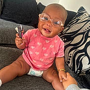 Ameileana joined the competition — help win amazing prizes! baby, glasses, pink_shirt, heart_pattern, doll, couch, cushion, hand_holding, smiling, indoor, diaper, bracelet, person, child, happy, seated, toy, face, casual_clothing, home