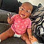 baby, glasses, pink_shirt, heart_pattern, doll, couch, cushion, hand_holding, smiling, indoor, diaper, bracelet, person, child, happy, seated, toy, face, casual_clothing, home