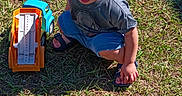 Elyis is registered to the contest to win money with this photo: blue_sippy_cup, child, crouching, curly_hair, daytime, expression, footwear, grass, greenery, nature, outdoor, plastic_toys, playing, sandals, shorts, sunlight, toddler, toy_trucks, tshirt, young_child