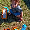 toddler, child, grass, toy_trucks, blue_sippy_cup, curly_hair, outdoor, sunlight, sandals, shorts, tshirt, playing, crouching, daytime, greenery, young_child, plastic_toys, footwear, expression, nature