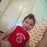 Elyis is registered to the contest to win money with this photo: toddler, child, smiling, red_shirt, indoor, home, quilt, pattern, door, wall, person, happy, sitting, curly_hair, casual_clothing, floor, light, portrait, young_child, cute