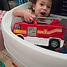 bath_time, bathroom, bathtub, child, childhood, curly_hair, floor_tiles, fun, happy, indoor, person, plastic_toys, playing, red_toy, smiling, toddler, toy_fire_truck, water_play, white_bathtub, young_child