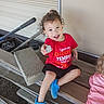 bench, blue_crocs, casual_clothing, child, concrete_block, curly_hair, fun, garden, happy, outdoor, person, playful, red_shirt, rock, sitting, smiling, toddler, tool, wooden_steps, young_child