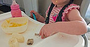 Alyiah is registered to the contest to win money with this photo: baby, banana, bottle, child, curly_hair, cute, eating, face, food, high_chair, indoor, kitchen, meal_time, person, pink, plastic_bowl, seat, snack, toddler, utensil