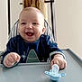 Maëcio participe au concours pour gagner de l'argent avec cette photo : baby, high_chair, pacifier, smiling, infant, blue_clothing, wood, neutral_background, happy, child, table, indoor, person, face, hand, cute, young, toddler, sitting, playful