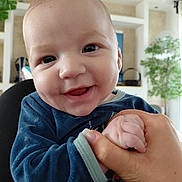 Maëcio a rejoint le concours — aidez-le/la à gagner de superbes lots ! baby, smile, hand, indoor, person, child, happy, face, blue_clothing, sleeve, holding, skin, fingers, adult_hand, background, plant, shelf, blurred_background, portrait, closeup