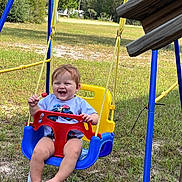 Carter is registered to the contest to win money with this photo: baby, child, swing, playground, outdoor, smiling, laughing, happy, lollipop, infant, sitting, play_equipment, blue, yellow, red, grass, backyard, trees, sunny, portrait