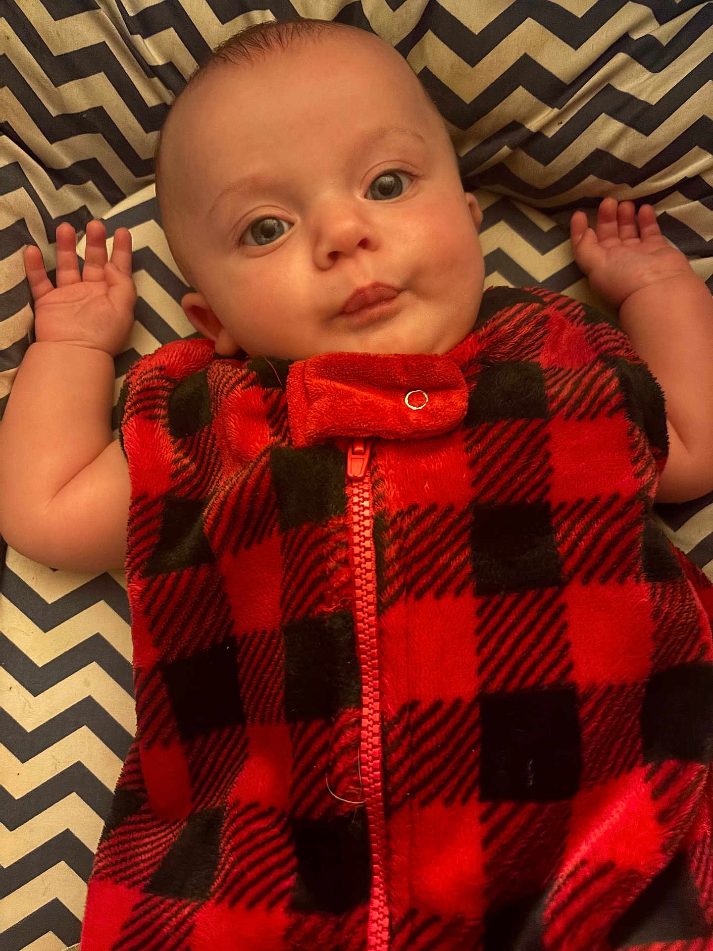 Jameson is registered to the contest to win money with this photo: baby, infant, face, eyes, hand, arm, clothing, fleece, red, black, plaid, pattern, blanket, zigzag, person, cute, lying_down, indoor, portrait, child