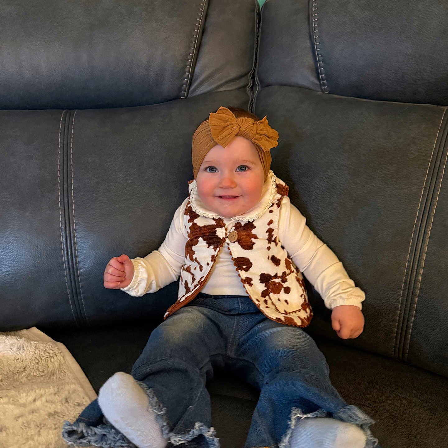 Maeleigh is registered to the contest to win money with this photo: baby, blanket, bow, child, couch, cow_print, cozy, denim, furniture, headband, infant, jeans, living_room, long_sleeve, portrait, sitting, smile, socks, sofa, vest