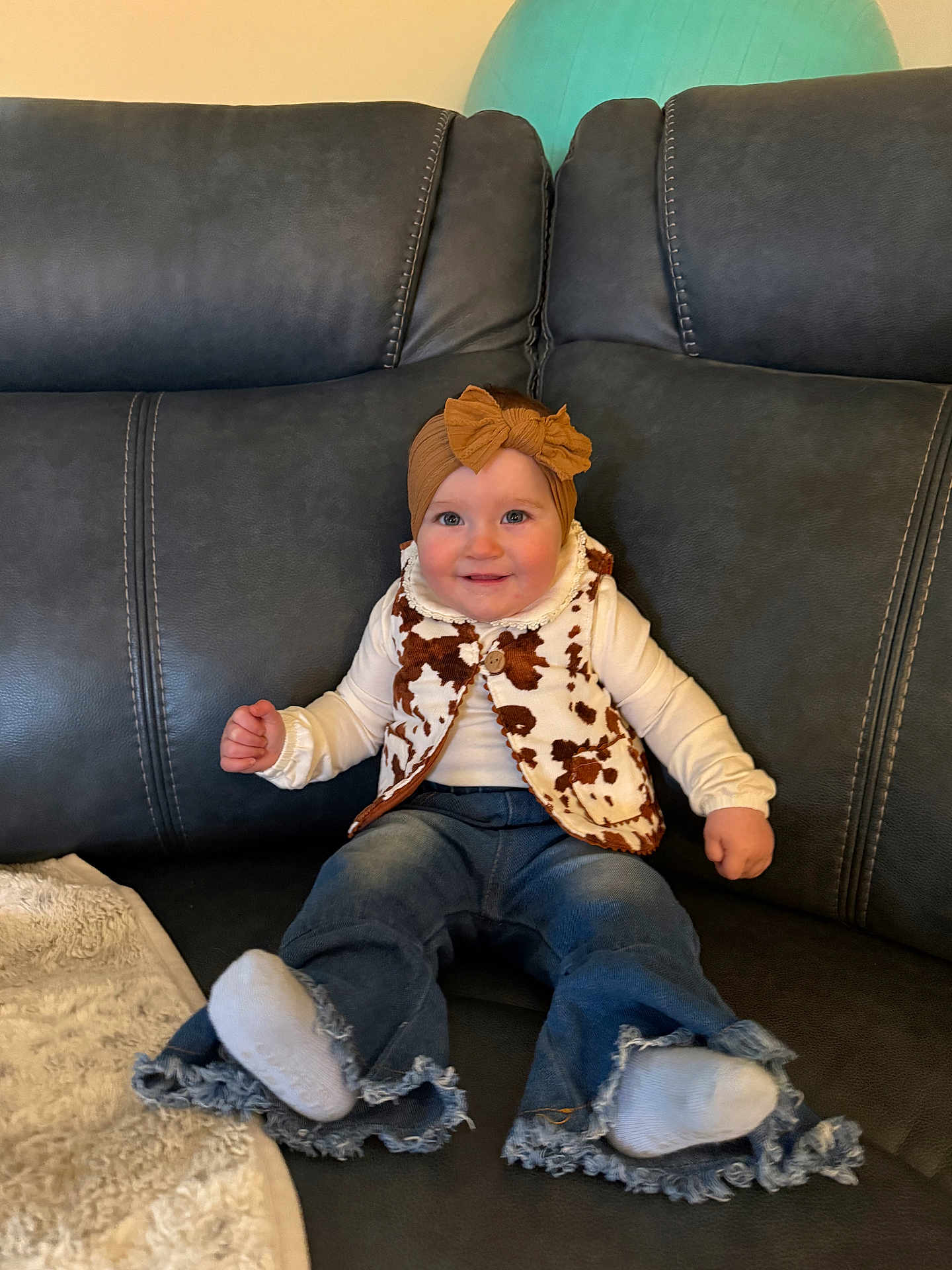 Maeleigh is registered to the contest to win money with this photo: baby, infant, child, couch, sofa, sitting, smile, headband, bow, vest, cow_print, long_sleeve, jeans, denim, socks, blanket, living_room, portrait, cozy, furniture