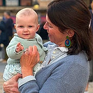 Céleste a rejoint le concours — aidez-le/la à gagner de superbes lots ! baby, woman, child, holding_child, smile, portrait, blue_sweater, knitwear, earrings, ring, hand, outdoor, urban, building, window, blurred_background, public_space, cute, close_up, daylight