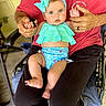 baby, child, adult, hands, blue_eyes, headband, swimsuit, sitting, lap, clothing, red_shirt, black_pants, indoor, chair, flooring, person, holding_hands, curious, cute, portrait