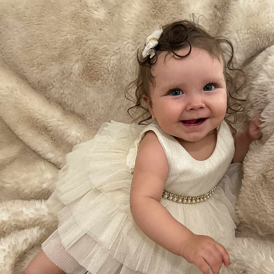 Adelina is registered to the contest to win money with this photo: baby, child, nine_months, dress, white_dress, smiling, hair_clip, curly_hair, fluffy_blanket, soft_texture, milestone, cute, infant, sitting, portrait, happy, indoor, toddler, young_child, celebration