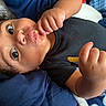 Zamir is registered to the contest to win money with this photo: baby, infant, child, portrait, lying_down, wide_eyed, curly_hair, smiling, snack, food, eating, hand, black_shirt, blanket, blue_background, close_up, face, eyes, cozy, indoor