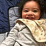 baby, child, infant, smiling, happy, portrait, curly_hair, brown_eyes, face, indoors, blanket, bib, gray_clothing, seated, baby_seat, cushion, bed, cozy, photography, close_up