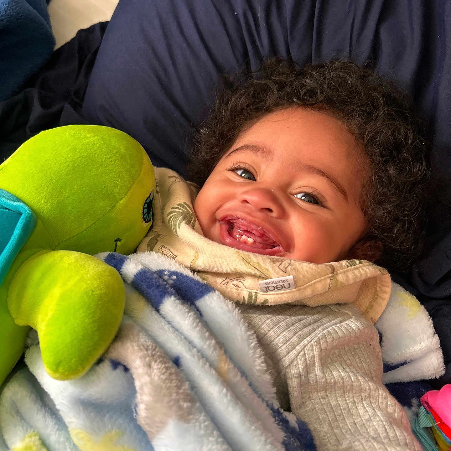Zamir joined the competition — help win amazing prizes! baby, child, infant, smiling, happy, portrait, face, curly_hair, eyes, teeth, blanket, plush_toy, stuffed_animal, bed, pillow, cozy, indoor, close_up, sitting, colorful