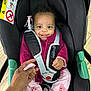 baby, child, car_seat, smiling, holding_hand, indoor, clothing, jacket, pants, socks, teddy_bear_patch, black_car_seat, safety_harness, adult_hand, wooden_floor, cute, portrait, infant, playful, seat_belt