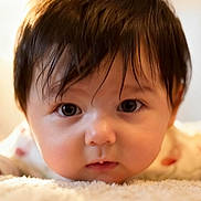 Mila is registered to the contest to win money with this photo: baby, close_up, face, child, infant, cute, dark_hair, eyes, skin, portrait, lying_down, soft_texture, warm_light, expression, young, adorable, focus, innocence, fluffy, head